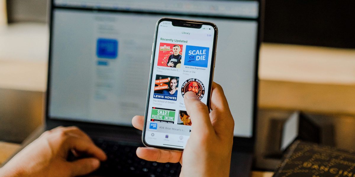 A person holds a smartphone displaying a Spotify report of podcasts, with a laptop in the background. The screen shows various podcast titles and colorful icons, creating a casual, tech-savvy atmosphere.