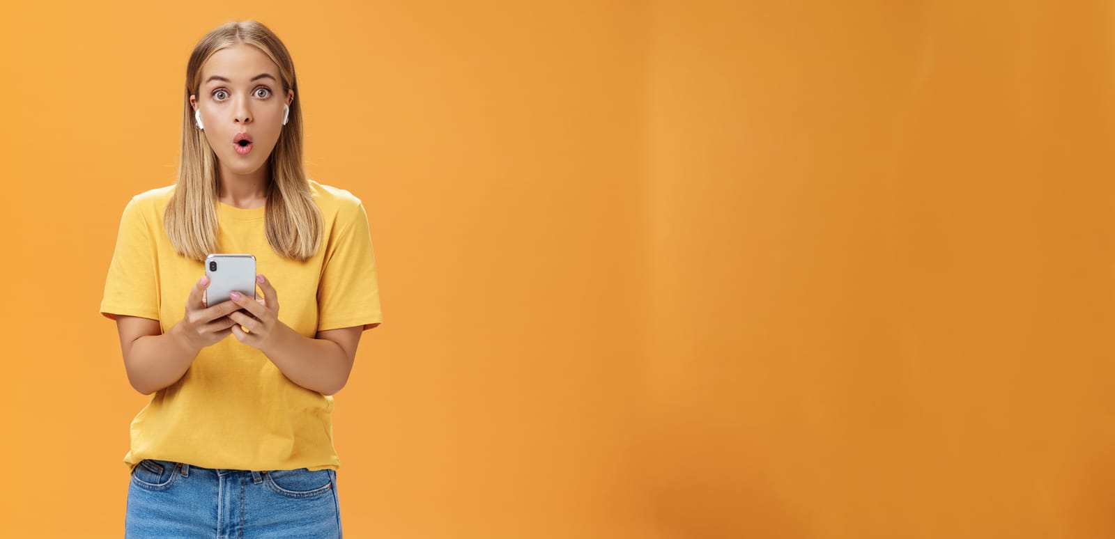 A woman wearing a yellow t-shirt and jeans holds a smartphone, wireless earbuds in her ears, displaying an expression of surprise. Against a solid orange background, she embodies the modern podcast listeners, fully immersed in captivating audio adventures.