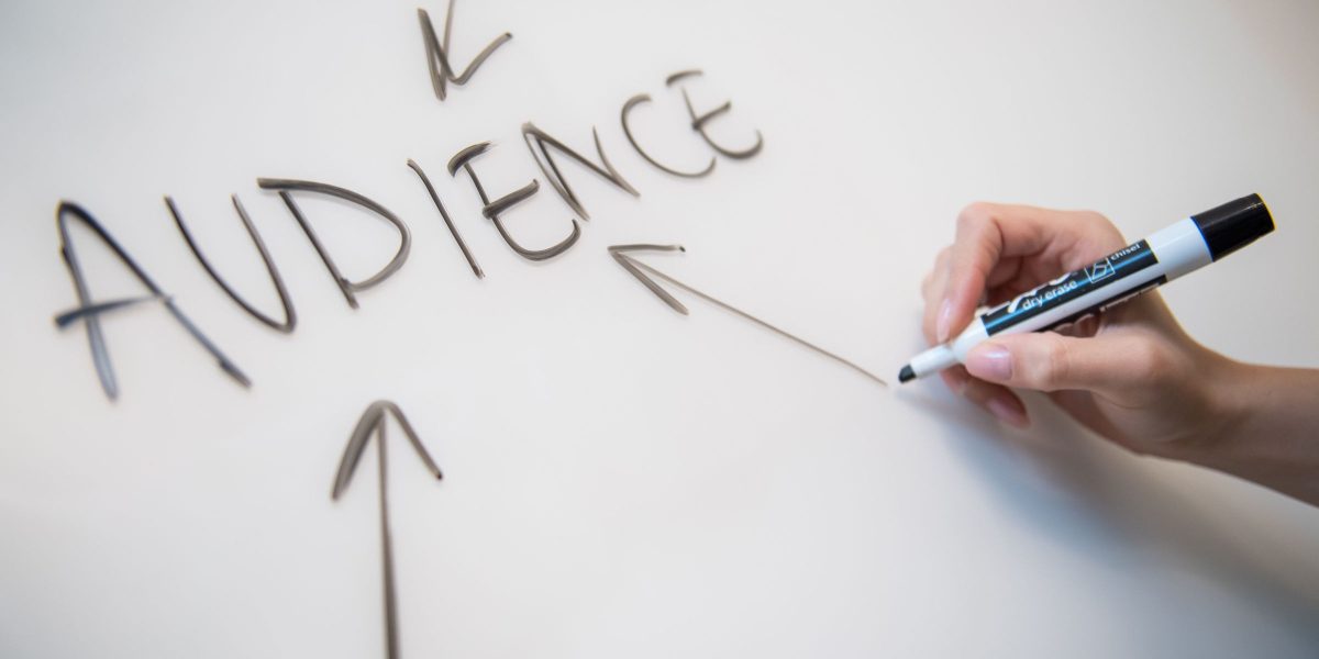 A hand skillfully writes AUDIENCE in large letters on a whiteboard, with arrows pointing toward the word from all directions. The person, holding a marker, strategizes ways of repurposing podcast content to engage and expand their reach.