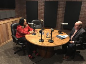 Two people sit at a round wooden table in a recording studio, engaging in a podcast interview. Both have microphones before them; the woman wears a red blouse, while the man is dressed in a suit. Their lively conversation fills the room with an unmistakable air of curiosity and inquiry.
