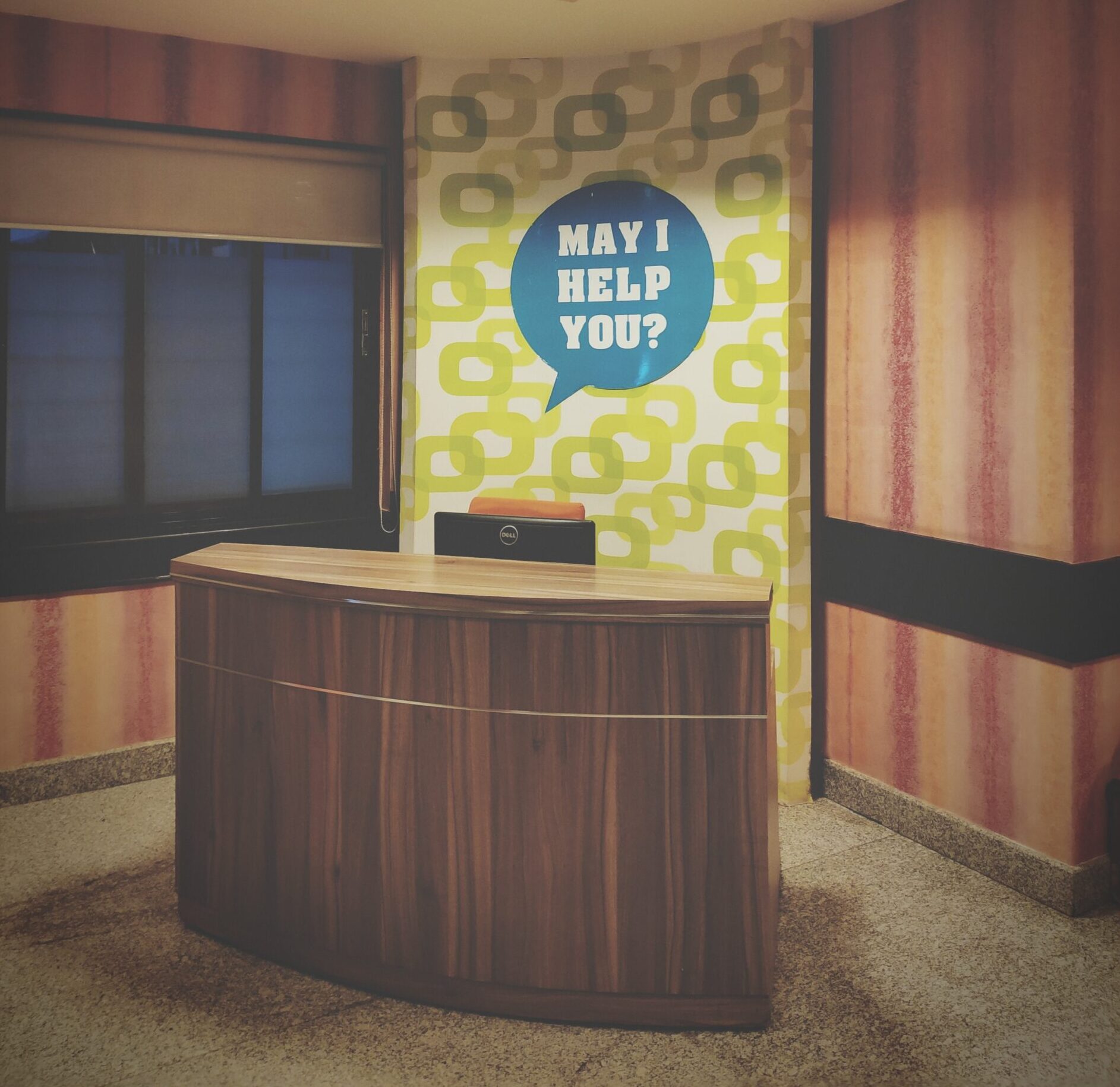 A wooden reception desk stands in front of a colorful wall with retro patterns, where a blue speech bubble reads, May I help you? Its the perfect creative space to launch your own podcast. The room features a window with a closed blind and speckled tiles underfoot.