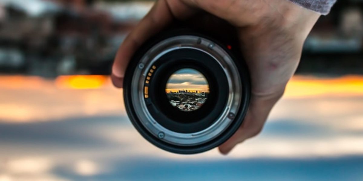 A person holds a camera lens, capturing an inverted reflection of a city skyline at sunset. Like finding a unique podcast niche, the scene through the lens is sharp and distinct, while the background is blurred, highlighting the colorful sky and urban silhouette.