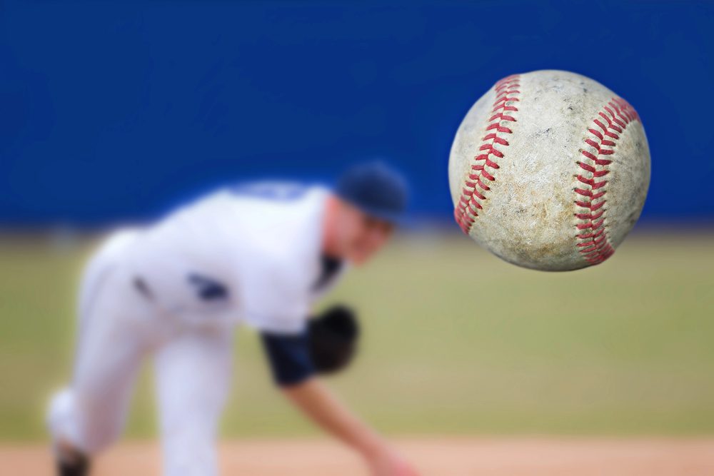 A baseball player in a white uniform is pitching a ball, much like a podcast guest catching the audiences attention. The focus is on the baseball in mid-air, with the player and field blurred in the background against a clear blue sky.