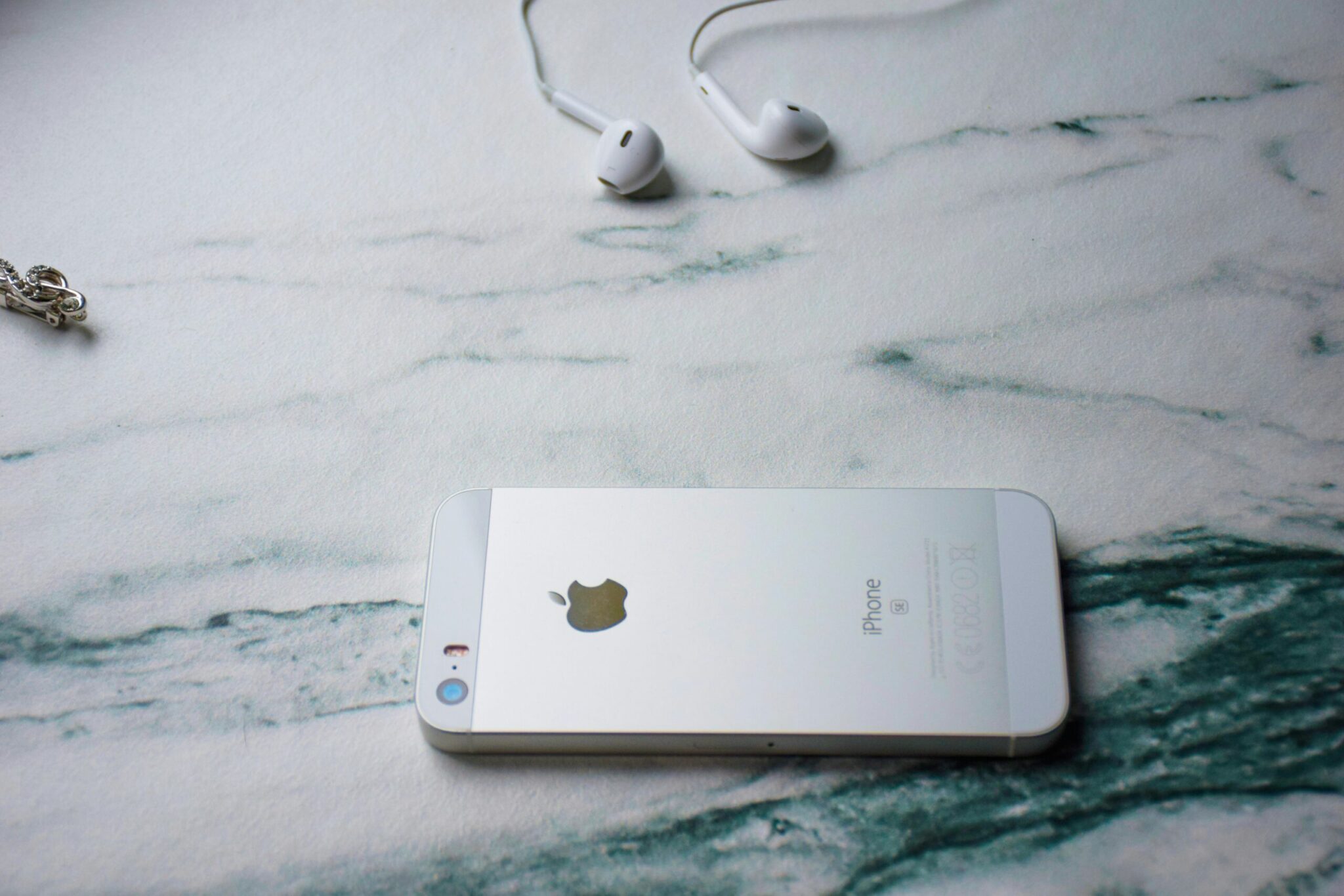 A silver iPhone lies face down on a marbled surface, ready to boost podcast engagement with its seamless connectivity, while a pair of white earbuds sit nearby. A delicate chain is partially visible in the corner, adding a touch of elegance to the scene.