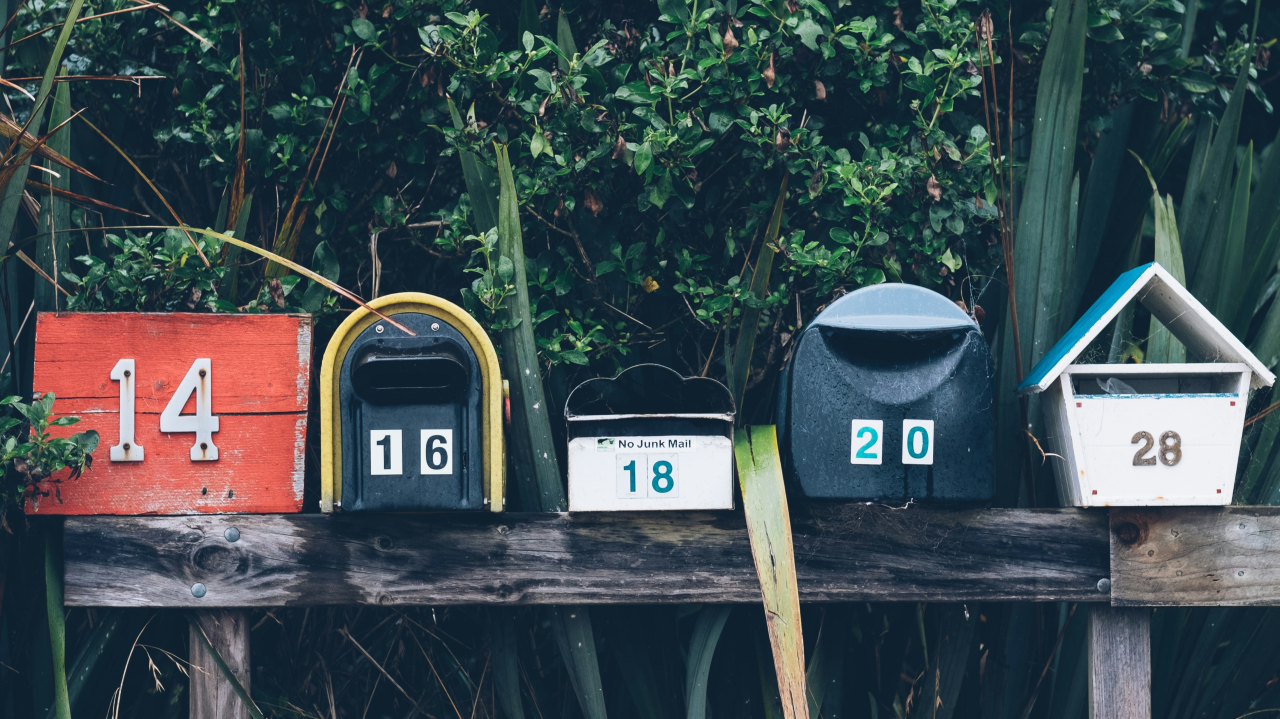Five mailboxes of different shapes and colors, numbered 14, 16, 18, 20, and 28, are mounted on a wooden post with green plants in the background—perfect for receiving cards or newsletters.