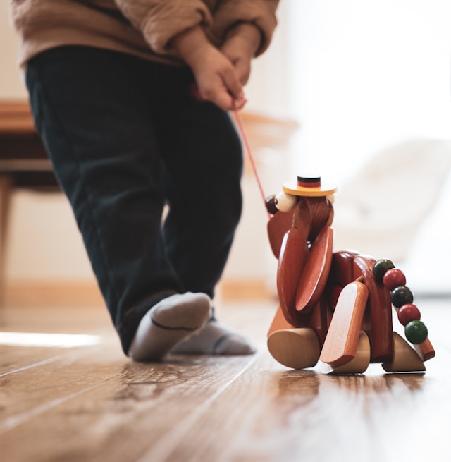 A small child in socks pulls a wooden toy animal with wheels and beads along a wooden floor, holding its string—much like how podcast audience engagement keeps listeners connected and moving forward. The child’s face is not visible.