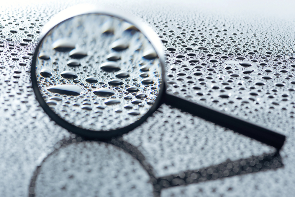 A magnifying glass lies on a wet surface with water droplets. The glass enlarges and distorts the droplets beneath it, revealing the power of reflection and creating a striking visual effect of varying droplet sizes.