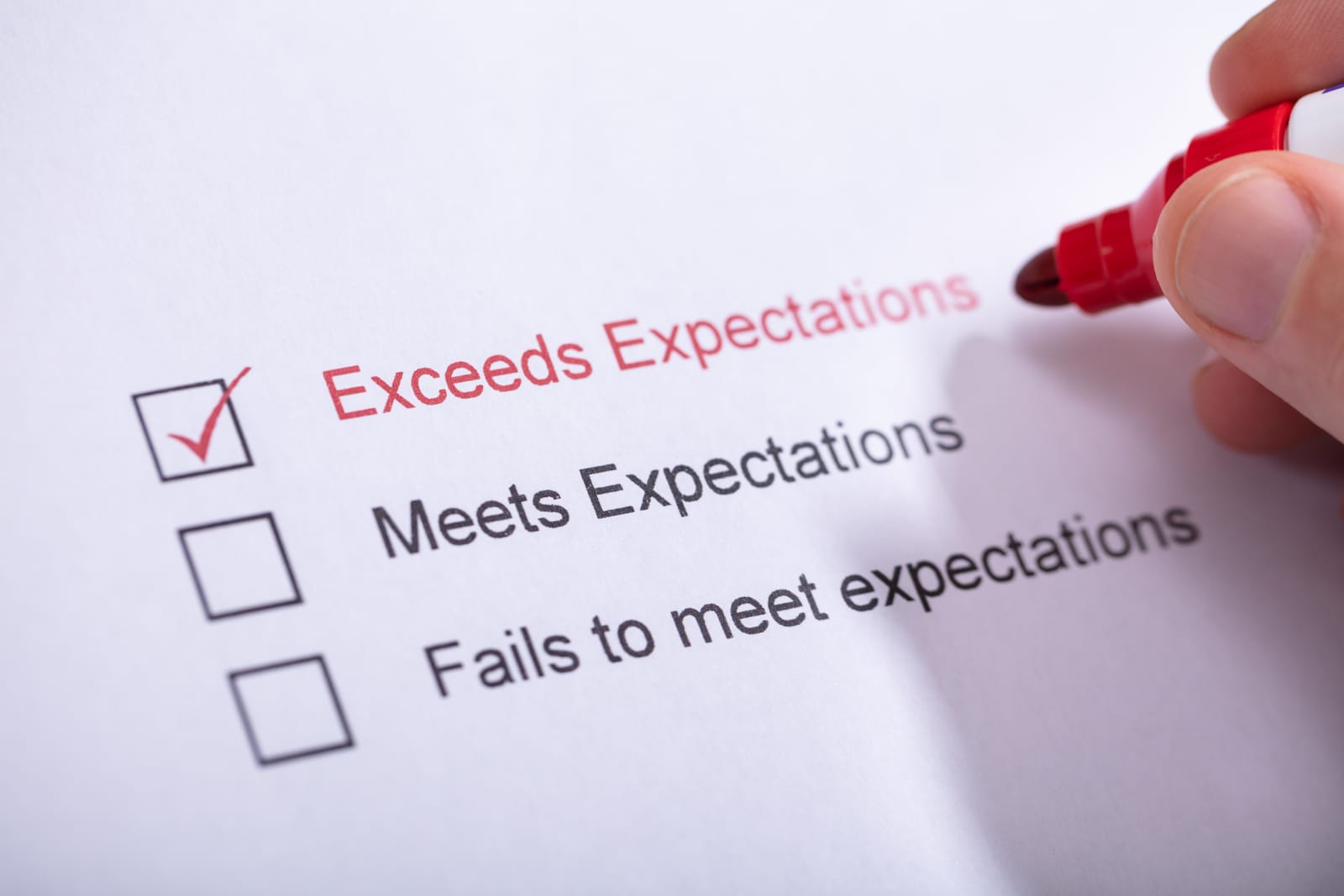A hand with a red marker checks the box next to Exceeds Expectations on a printed evaluation form, setting the standard for podcast expectations while the options Meets Expectations and Fails to meet expectations remain unchecked.