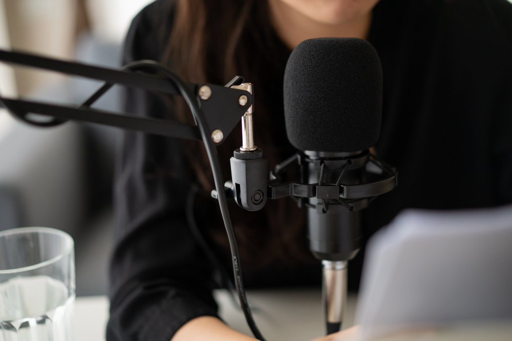 A person in black sits in front of a large microphone and pop filter, with a sheet of paper and a glass of water nearby, suggesting they are working on a creative project like recording a podcast or voiceover.