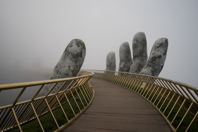 A curved wooden walkway with gold railings crosses through dense fog, flanked by giant stone hands that appear to support the bridge—a scene as mysterious and atmospheric as building a podcast email list for effective podcast marketing.