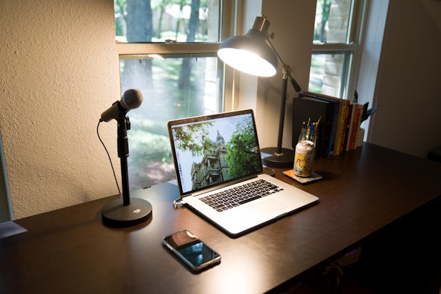 A laptop, microphone, smartphone, lamp, books, and a jar of pens are arranged on a dark wooden desk by a window with trees visible outside—an inviting setup that’s perfect for anyone looking to restart a podcast after a break.