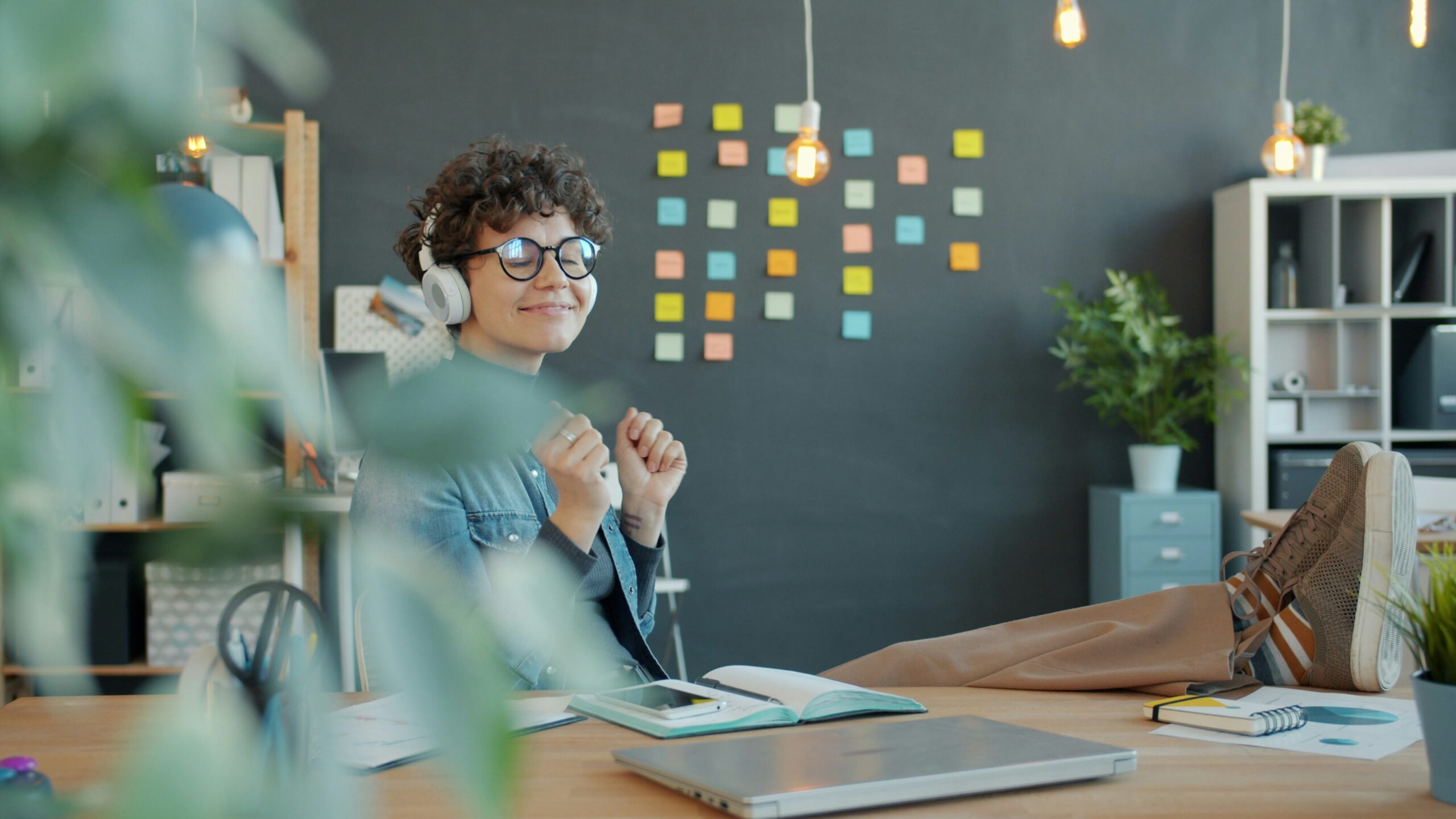 A person with curly hair and glasses, wearing headphones, sits at a desk with their feet up, smiling with eyes closed as if enjoying a favorite podcast. Colorful sticky notes cover a black wall in the background—perhaps reminders for an upcoming review or reasons why the workspace feels inspiring. Plants and office supplies are visible.