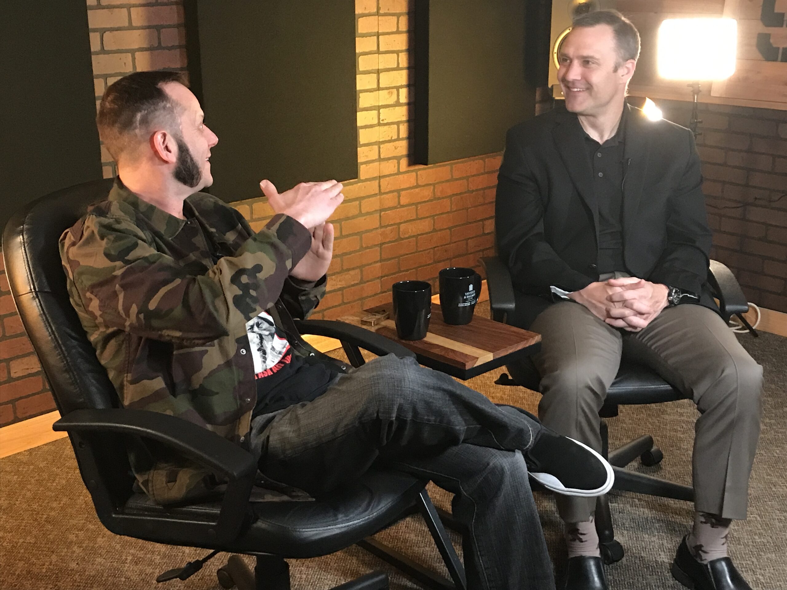 Two men sit and talk in a professional studio with brick walls. One gestures while speaking, wearing a camo jacket, and the other, in a suit, listens and smiles. Two black mugs sit on a small table between them, giving the space an inviting podcast atmosphere.